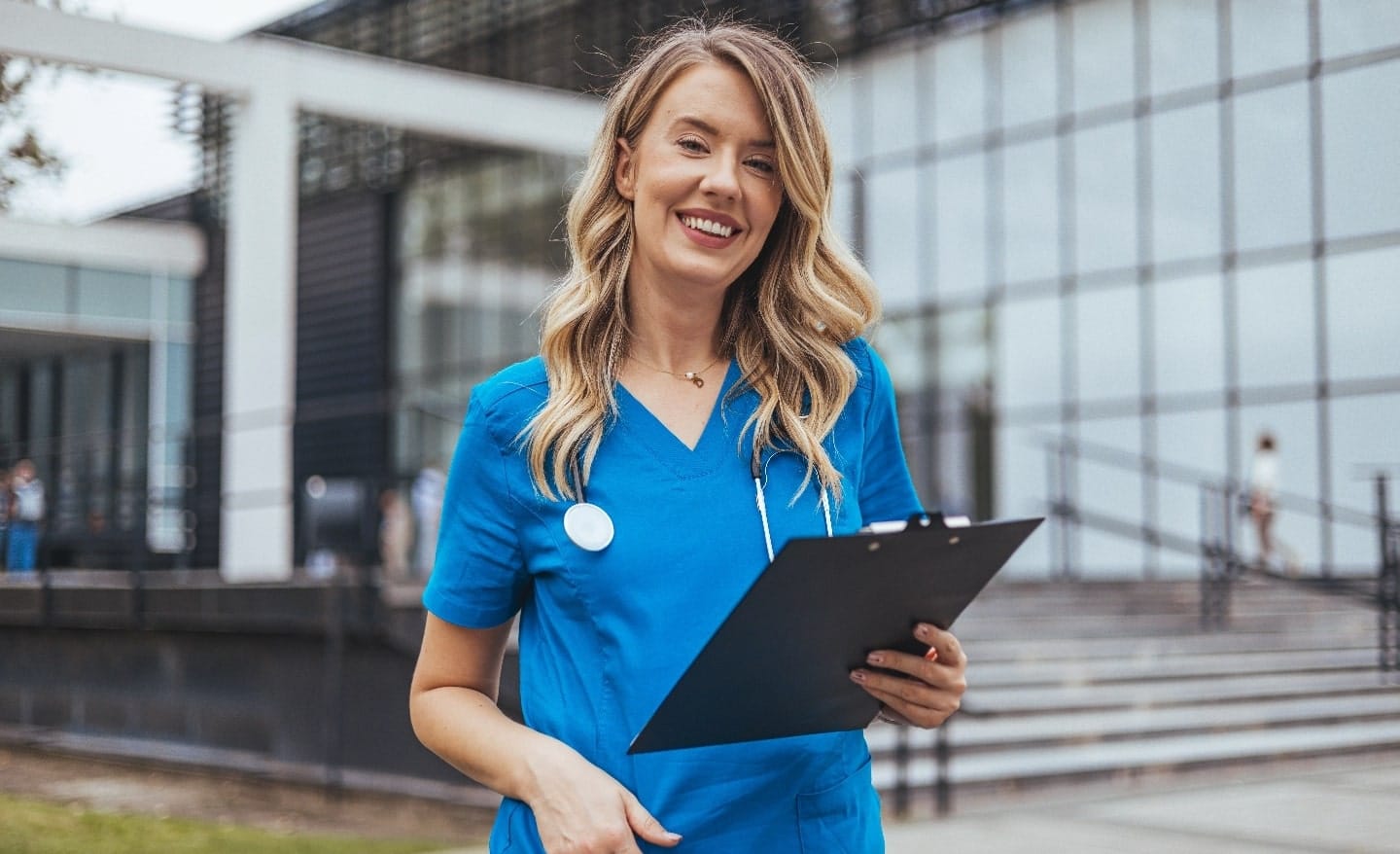 Smiling nurse in blue scrubs holding a clipboard outside a modern hospital building.