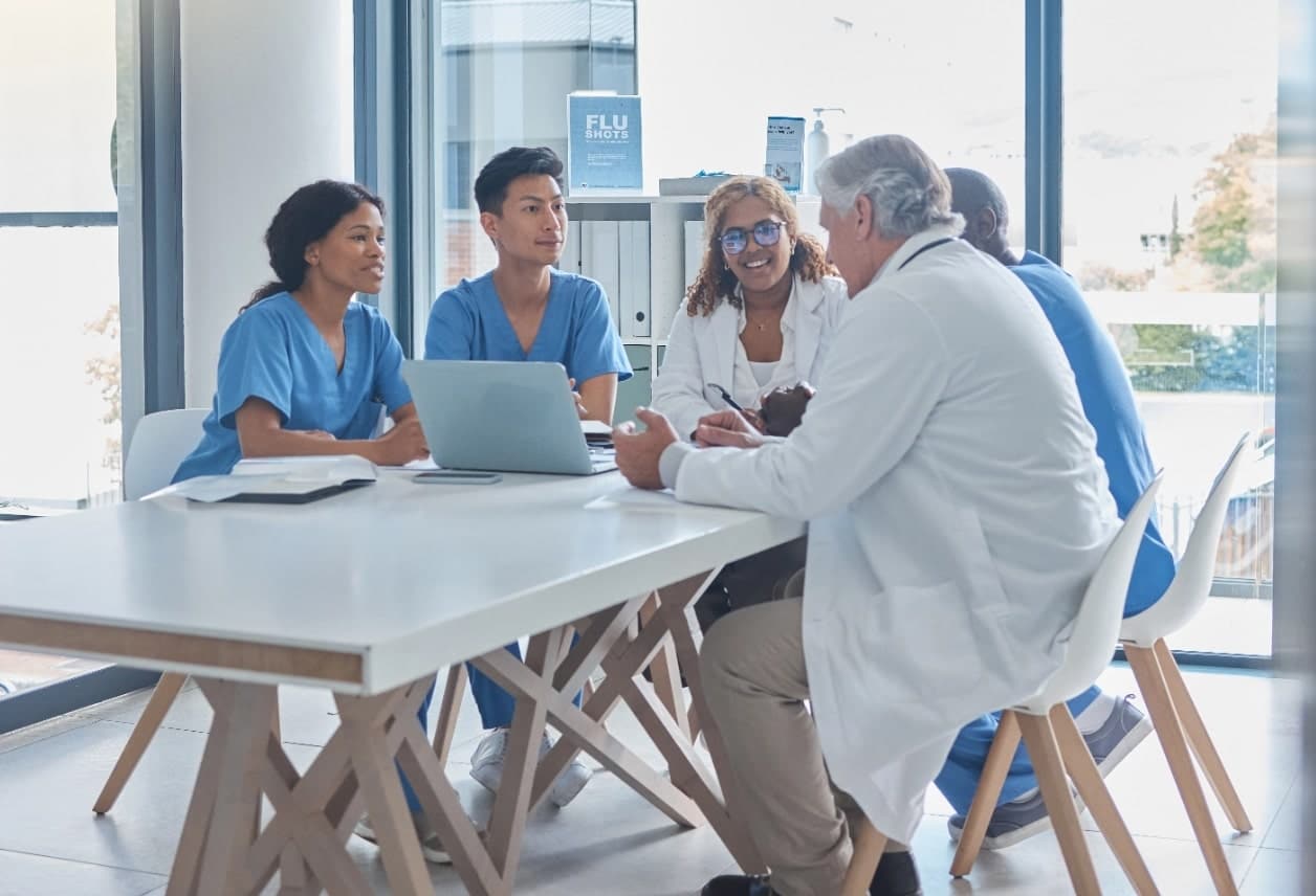 Healthcare team meeting around a table with laptop and notes.