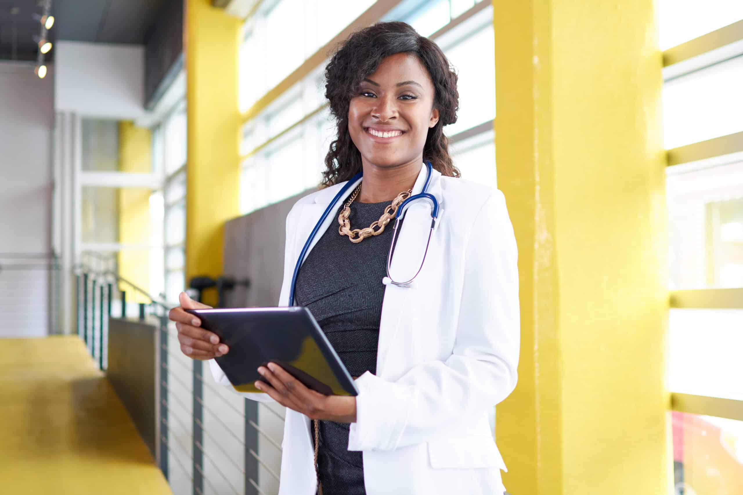 Smiling Black female doctor in white coat holding tablet. Stethoscope around neck.
